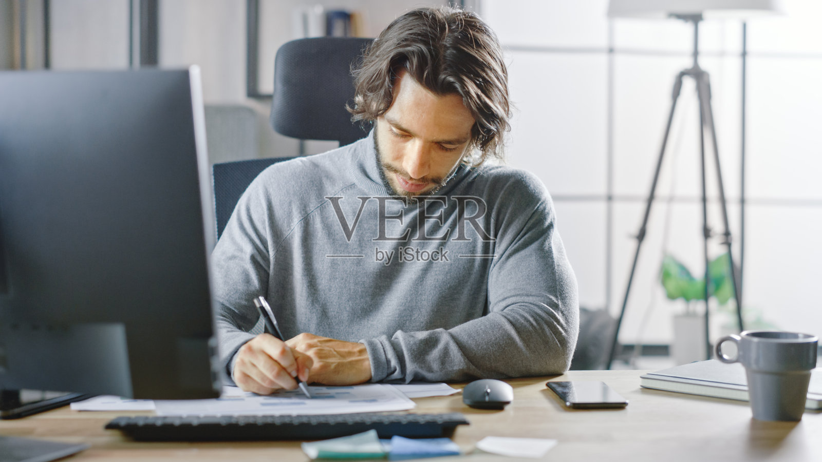 英俊的长发企业家坐在他的办公桌前，在台式电脑上工作，处理文件，图表，图表，统计和公司战略。商人填充文档照片摄影图片