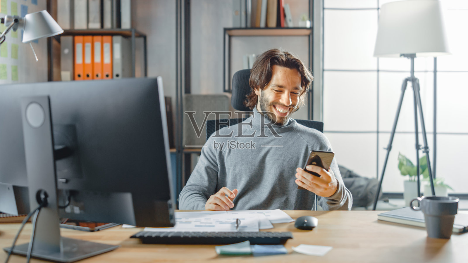 英俊的长发企业家坐在他的办公桌前，在台式电脑上工作，处理文件，图表。微笑使用智能手机，社交媒体应用程序，写电子邮件，短信照片摄影图片