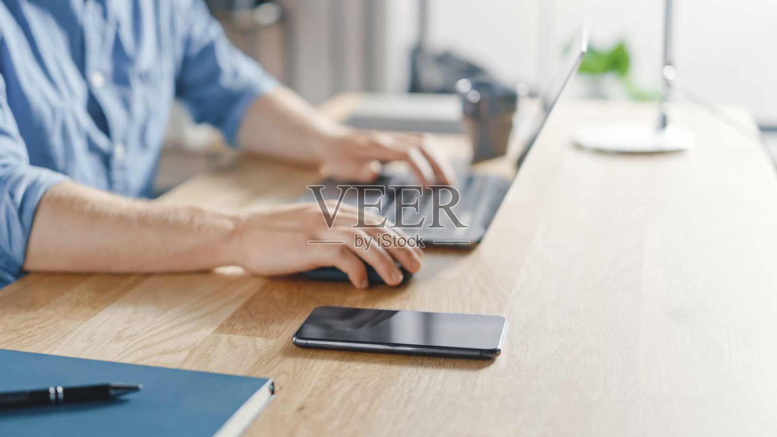 一个商人坐在他的办公桌前在时髦的办公室里用台式电脑工作的特写。他的智能手机就在身边。照片摄影图片