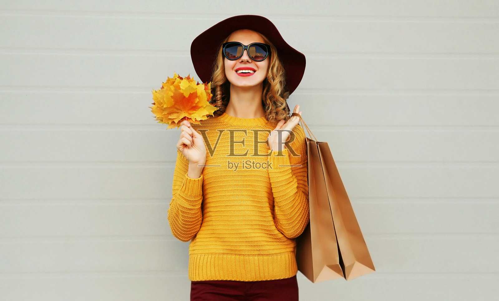 秋天的肖像女子拿着购物袋和黄色的枫叶穿着毛衣，帽子在灰色的背景照片摄影图片