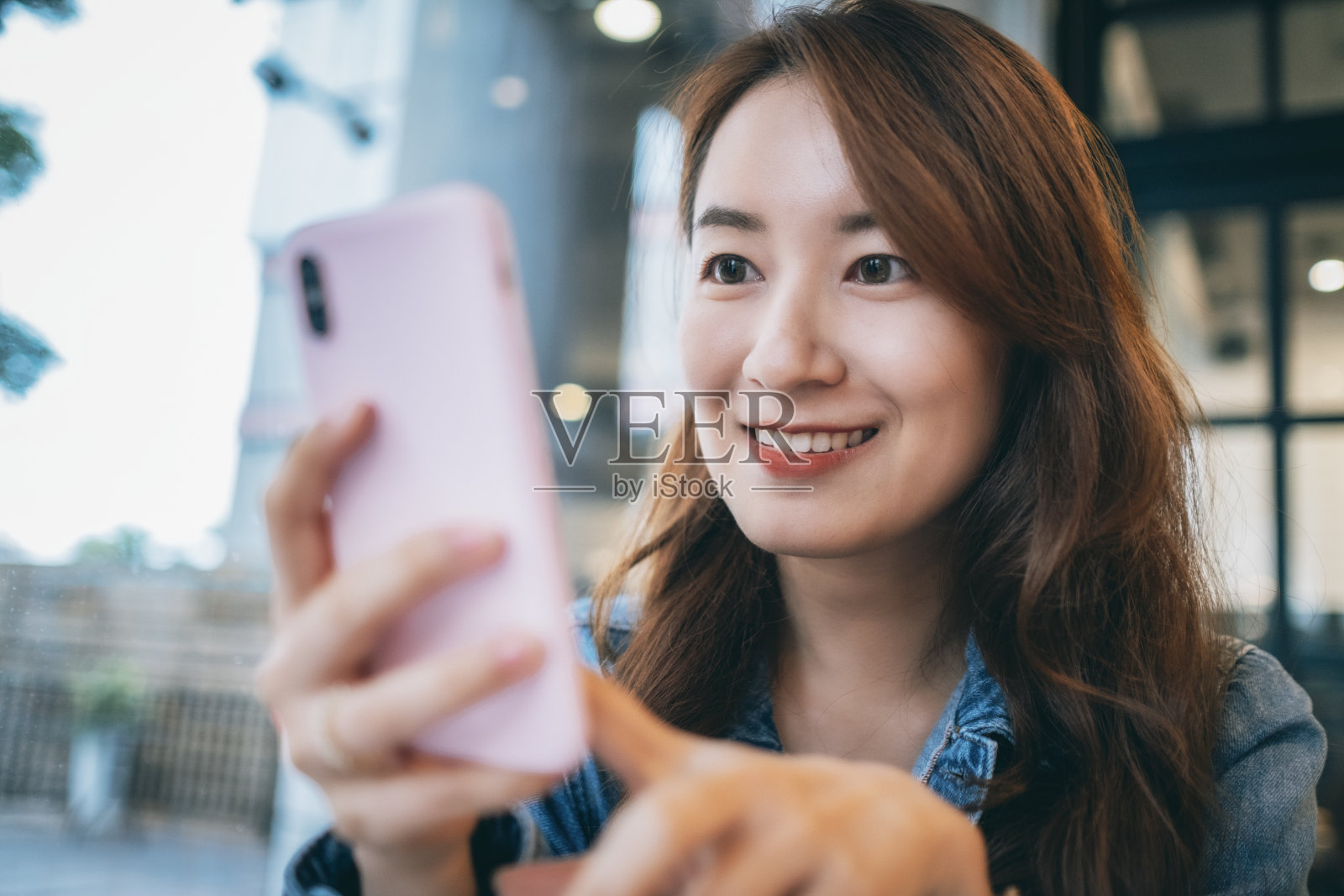 微笑的漂亮的年轻女子在休闲衬衫坐在桌子上的咖啡馆和查看短信，而使用智能手机照片摄影图片