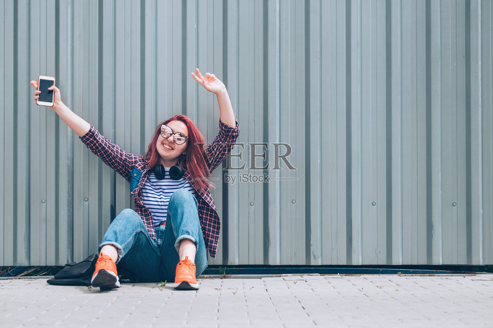 年轻美丽的现代微笑女性青少年，穿着格子衬衫和牛仔裤，戴着耳机和智能手机坐在街道的墙边，高兴的手举起。无忧无虑的学生年龄概念照片摄影图片