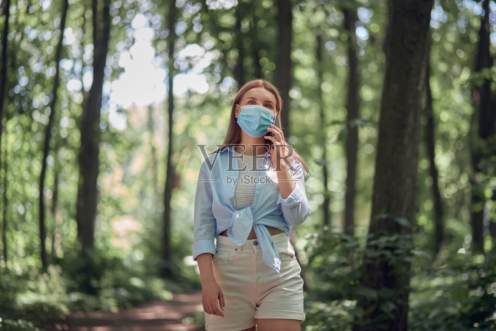 一个女孩戴着口罩走在公园里，用手机聊天。照片摄影图片
