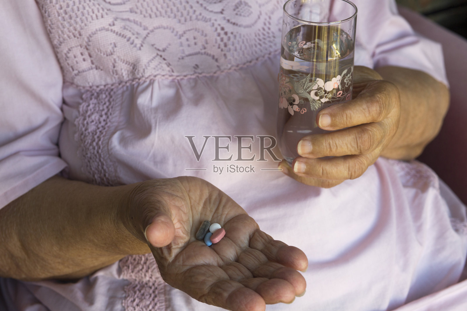 老妇人皱巴巴的手拿着一杯水和五颜六色的药丸。水平的特写。照片摄影图片
