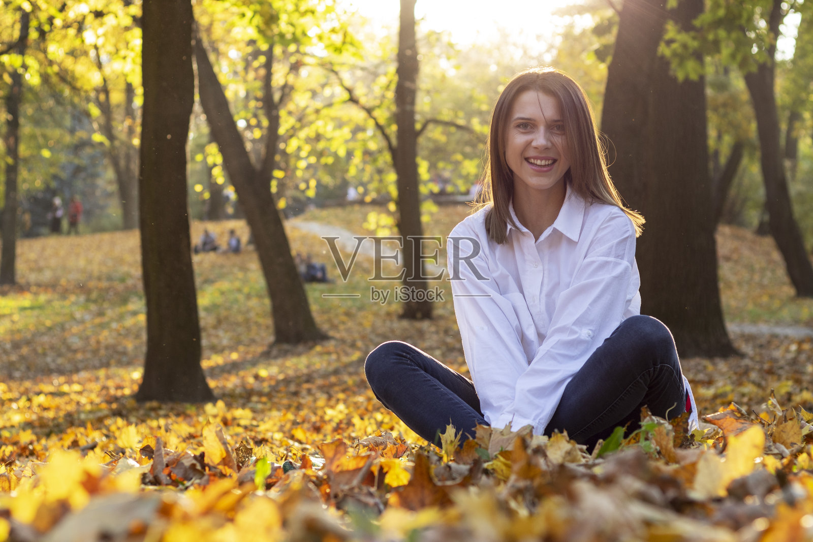 年轻微笑的女子坐在秋天公园的落叶上。照片摄影图片