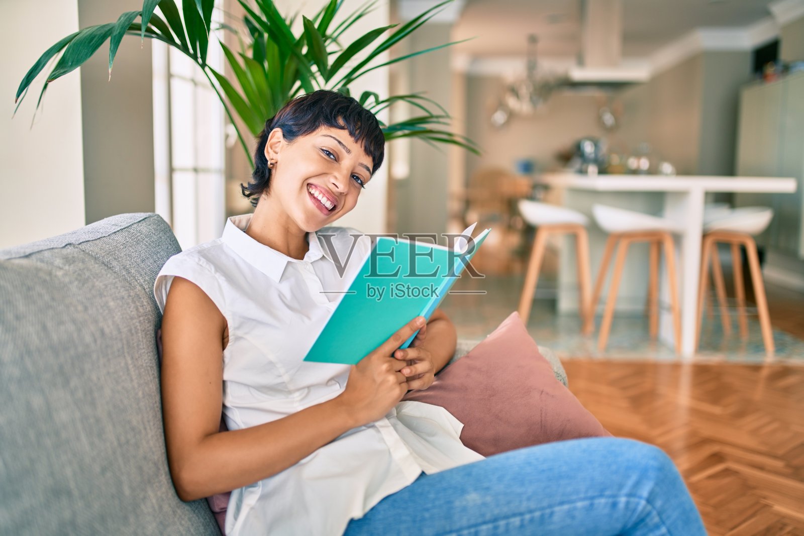 漂亮的黑发短发女人坐在家里的沙发上看书照片摄影图片