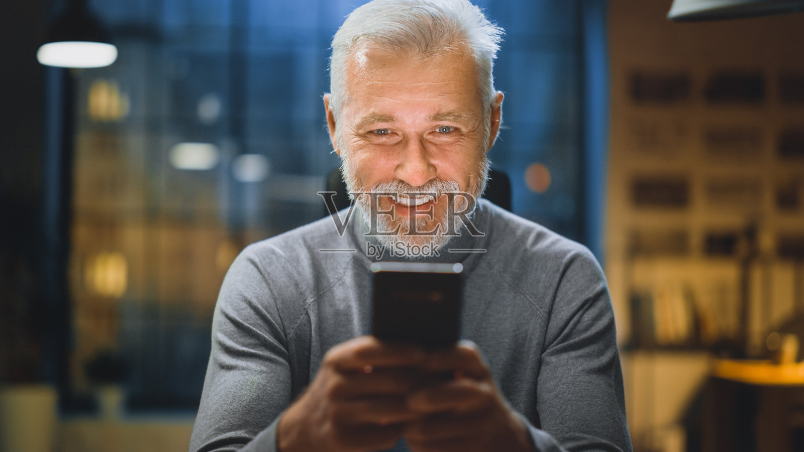 英俊和成功的中年胡子商人的肖像坐在他的办公桌上使用智能手机，大笑和微笑。在舒适的家庭办公室/工作室工作照片摄影图片