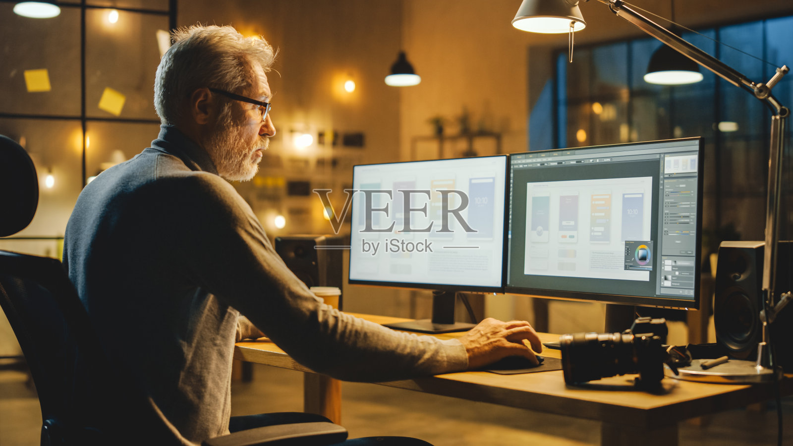 创意中年手机软件设计师坐在他的办公桌上使用桌面电脑和两个屏幕显示智能手机应用程序设计过程。时尚现代工作室办公室照片摄影图片