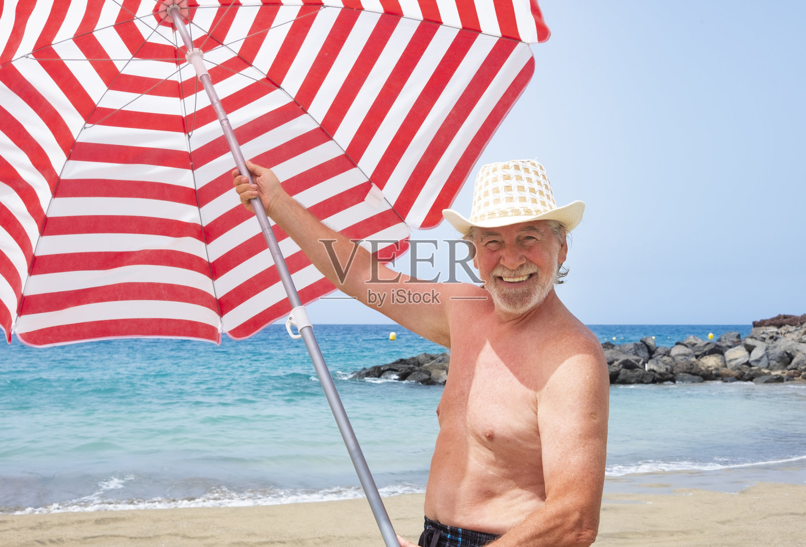 有魅力的大胡子老人戴着草帽在海滩上享受假期，拿着一把条纹伞，穿着泳衣和大大的微笑-活跃的退休老人和有趣的概念照片摄影图片