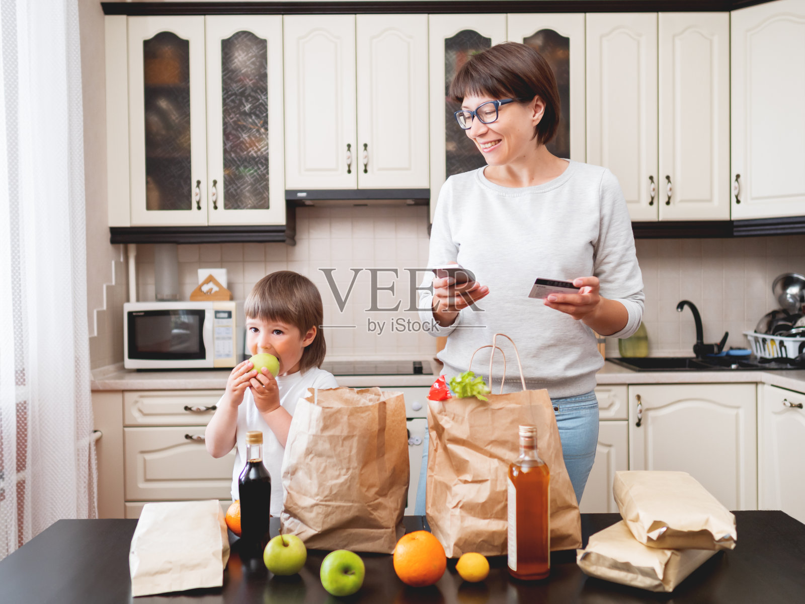 女人和蹒跚学步的男孩在厨房里挑选购买的东西。一个小孩咬了苹果。纸袋食品杂货配送。杂货店订阅服务。母亲和儿子在厨房。照片摄影图片
