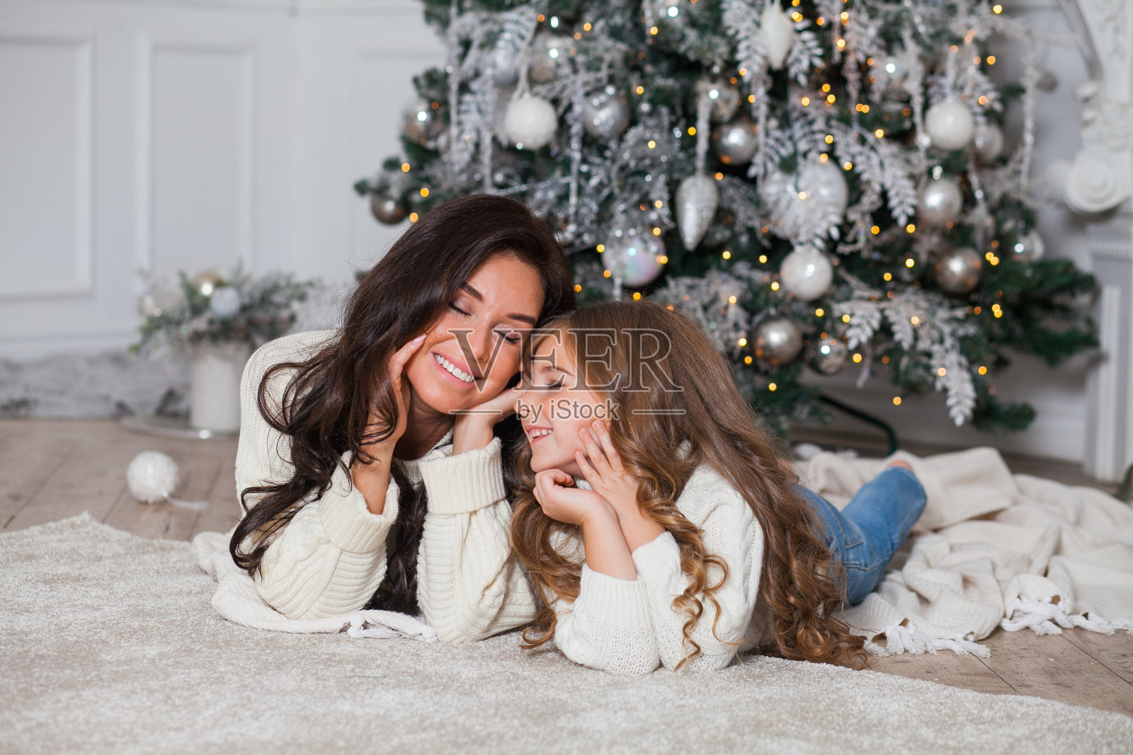 除夕合家欢乐。年轻美丽的母亲和小可爱的女儿在白色舒适的毛衣和毯子有乐趣和拥抱下装饰的圣诞树。节日家居经典室内设计照片摄影图片