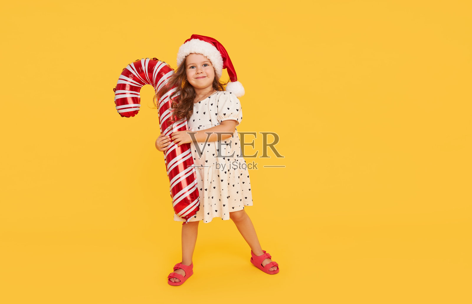 一个可爱的小女孩穿着漂亮的裙子，戴着圣诞老人的帽子，在黄色的背景上拿着一个充气的拐杖糖。2021年元旦假期概念及儿童新年礼品广告照片摄影图片