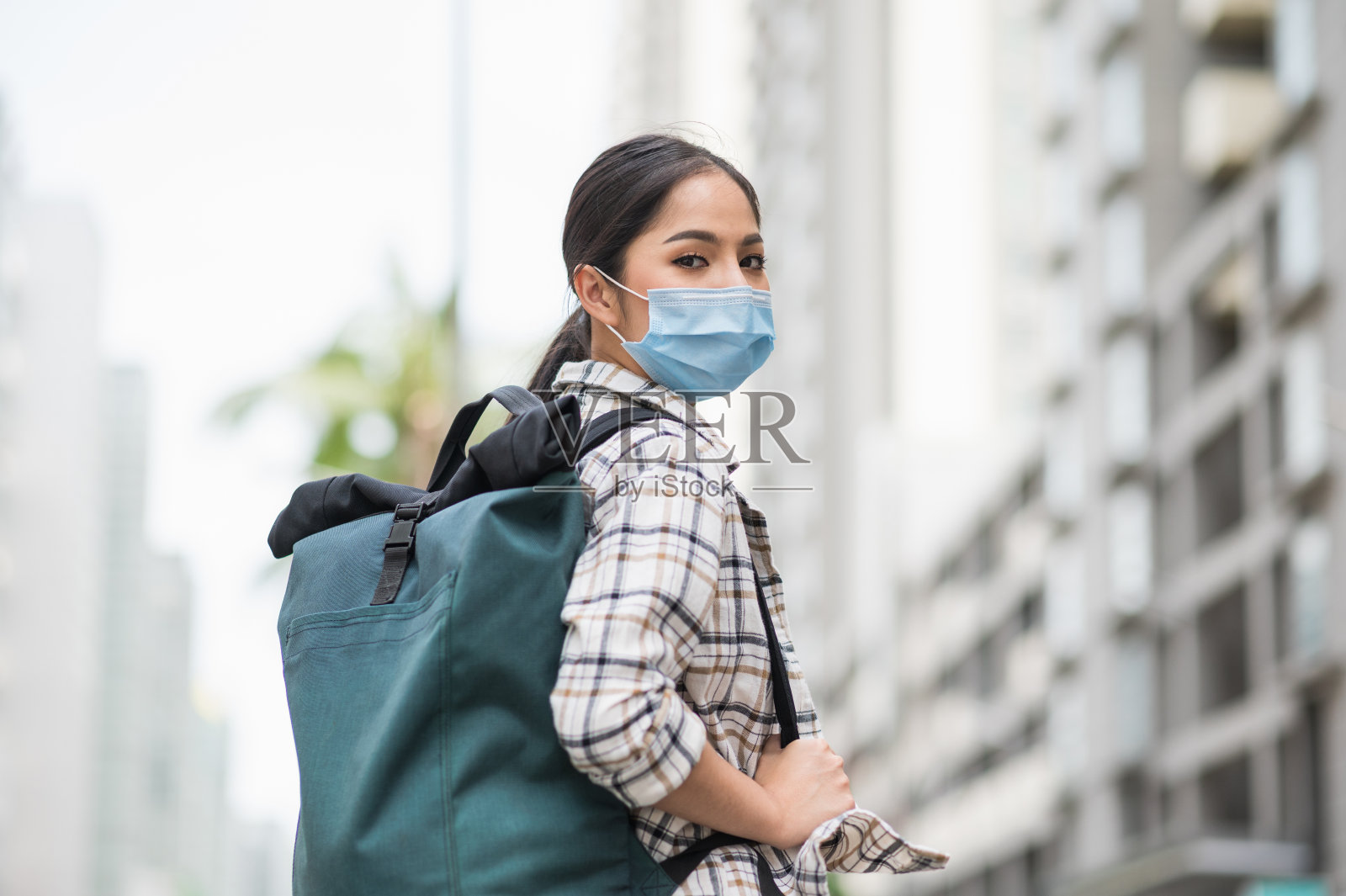 旅行者的新常态。孤芳自赏的年轻亚洲美女在旅行中背着背包，戴着防护口罩，安全旅行。她独自走在城市里。照片摄影图片