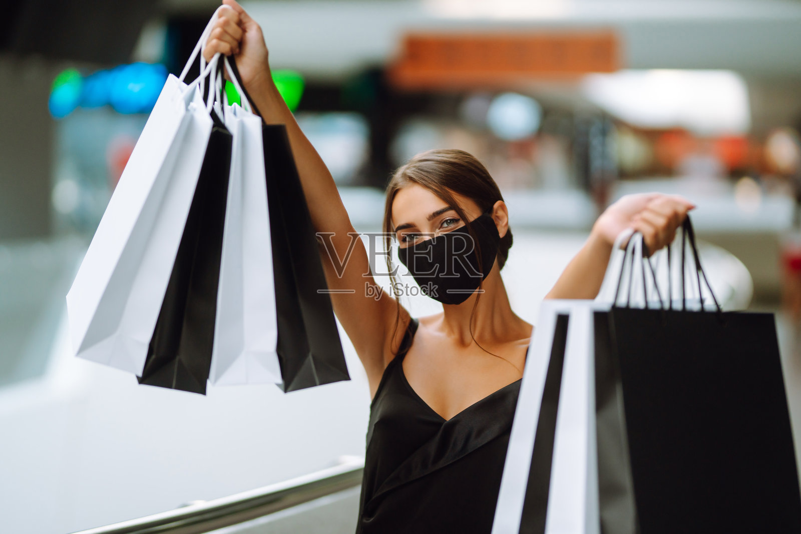 年轻女子防护黑色医用口罩在她的脸上与购物袋在商场。照片摄影图片