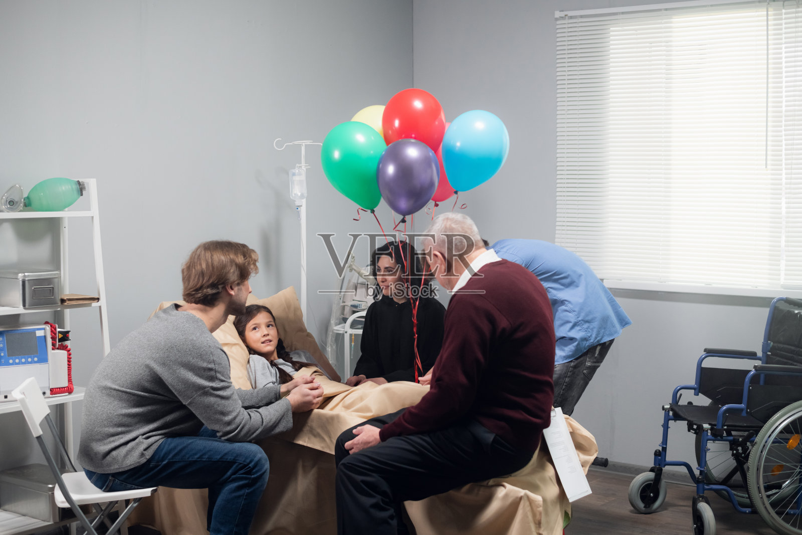 一个家庭去看望一个漂亮的小女孩，小女孩在一次事故后，躺在医院病房的床上，她的轮椅在角落里照片摄影图片