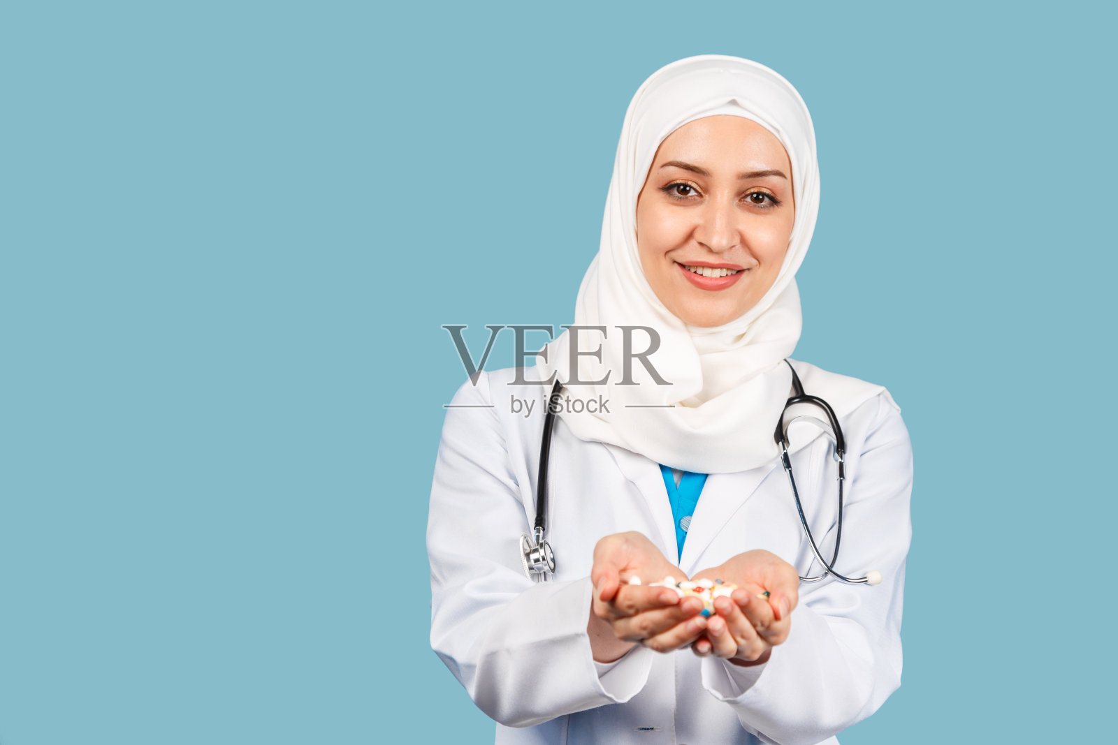 一名友好的穆斯林女医生的肖像，戴着头巾，手持听诊器，在蓝色背景下拿着一把药丸。照片摄影图片