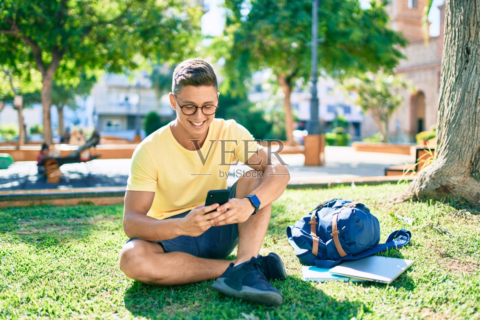 年轻的西班牙裔学生坐在大学校园的草地上，用智能手机开心地微笑着照片摄影图片