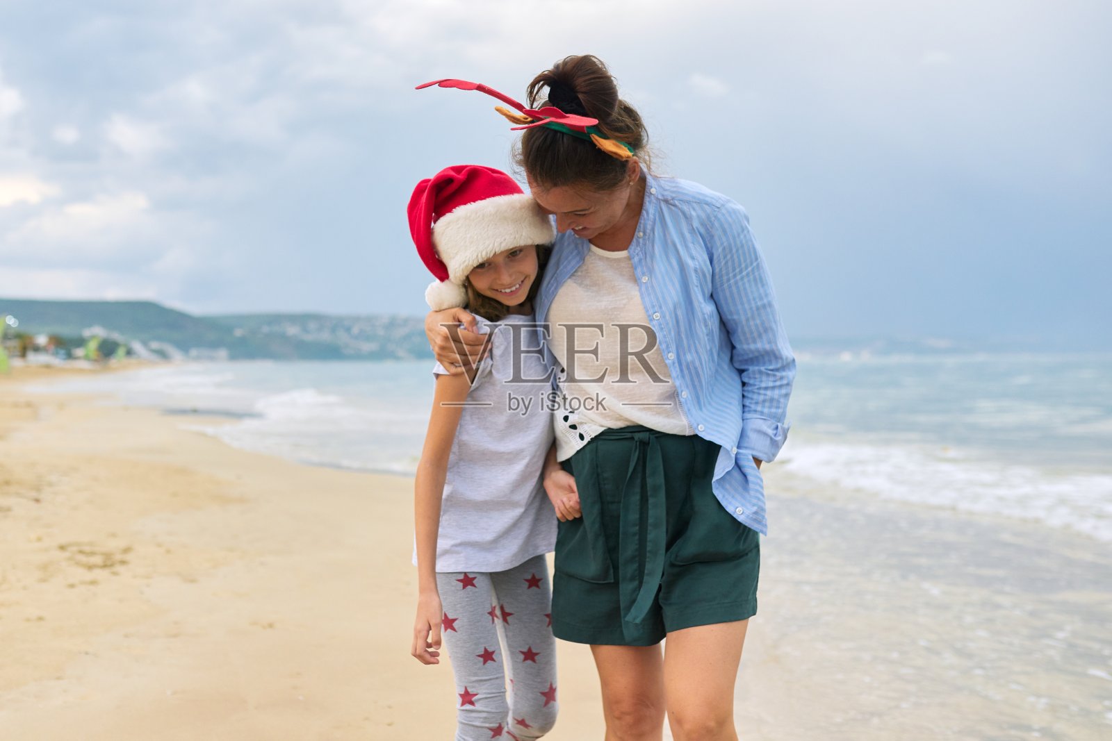 快乐的妈妈和女儿孩子在海滩上拥抱圣诞老人的帽子照片摄影图片