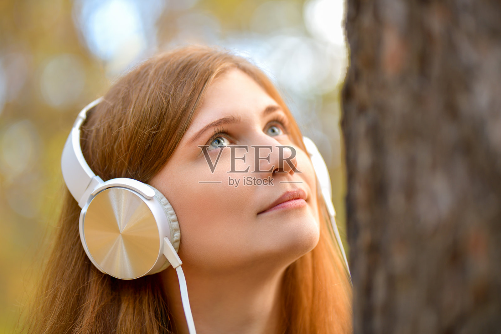 肖像。可爱的红发女孩戴着白色耳机看着树林里的一棵树。树林里戴耳机的女孩。女人看着那棵树。照片摄影图片