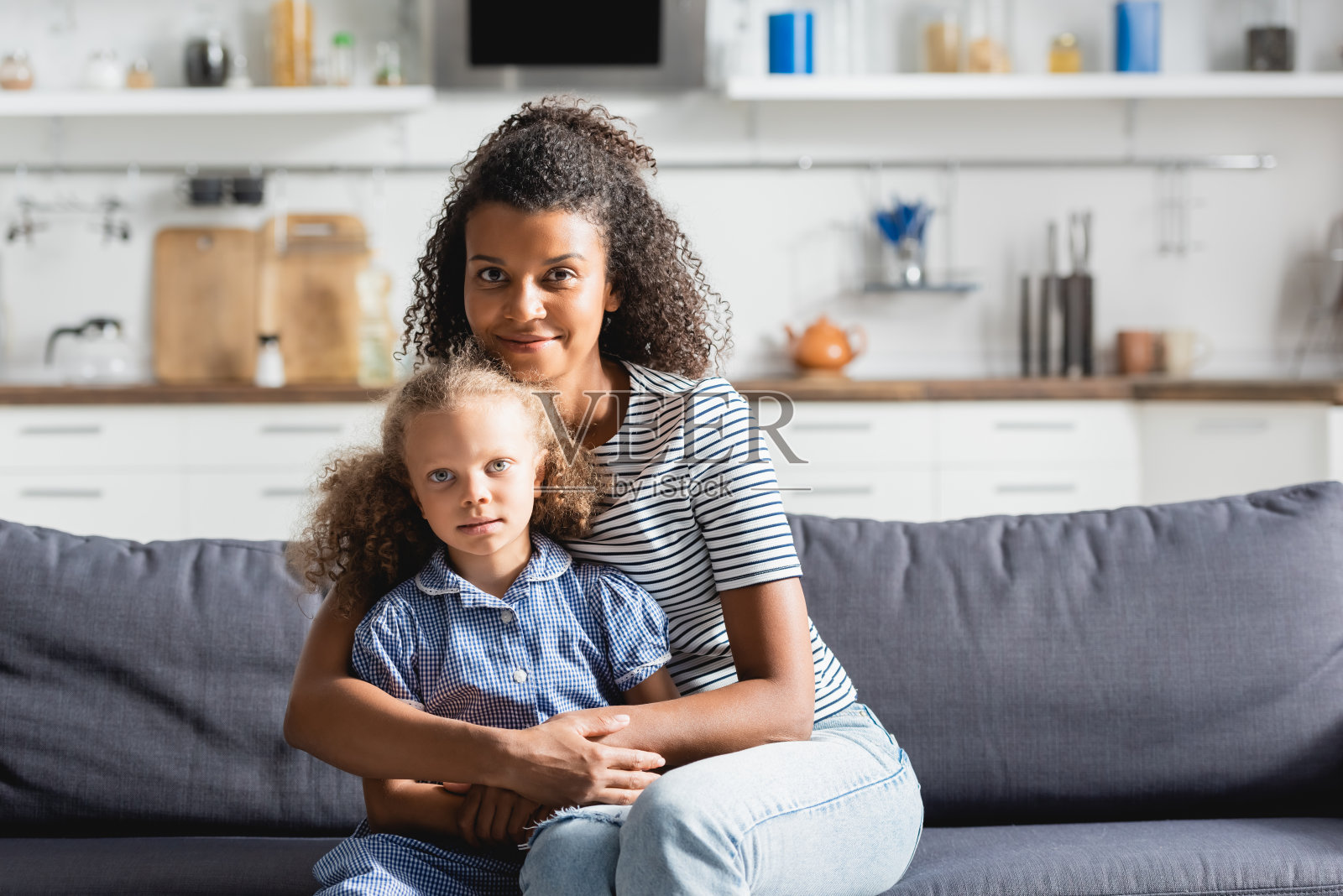 年轻的非洲裔美国妇女抱着女儿，坐在厨房的沙发上看着镜头照片摄影图片