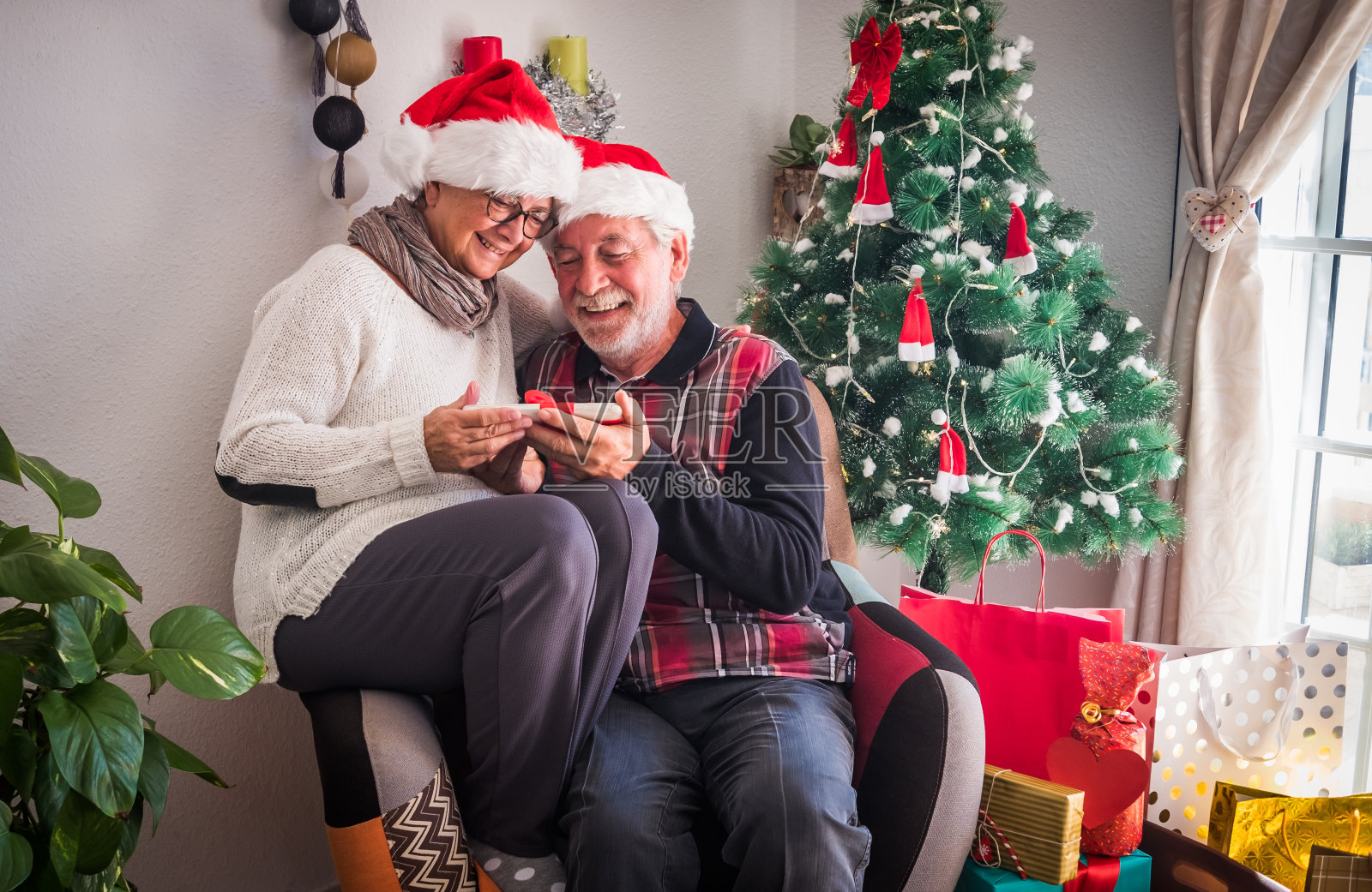 一对戴着圣诞老人帽的老人坐在同一张扶手椅上。拿着作为圣诞礼物收到的写字板。礼物和圣诞树在背景照片摄影图片