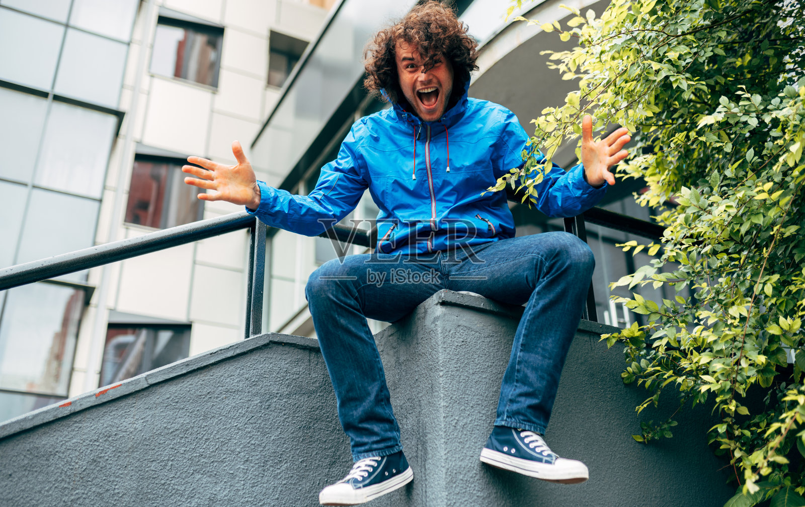 户外形象的一个愉快的年轻人卷曲的头发在蓝色雨衣坐在楼梯上举起手臂。一个下雨天在街上休息的男人。潮男有快乐的情绪。照片摄影图片