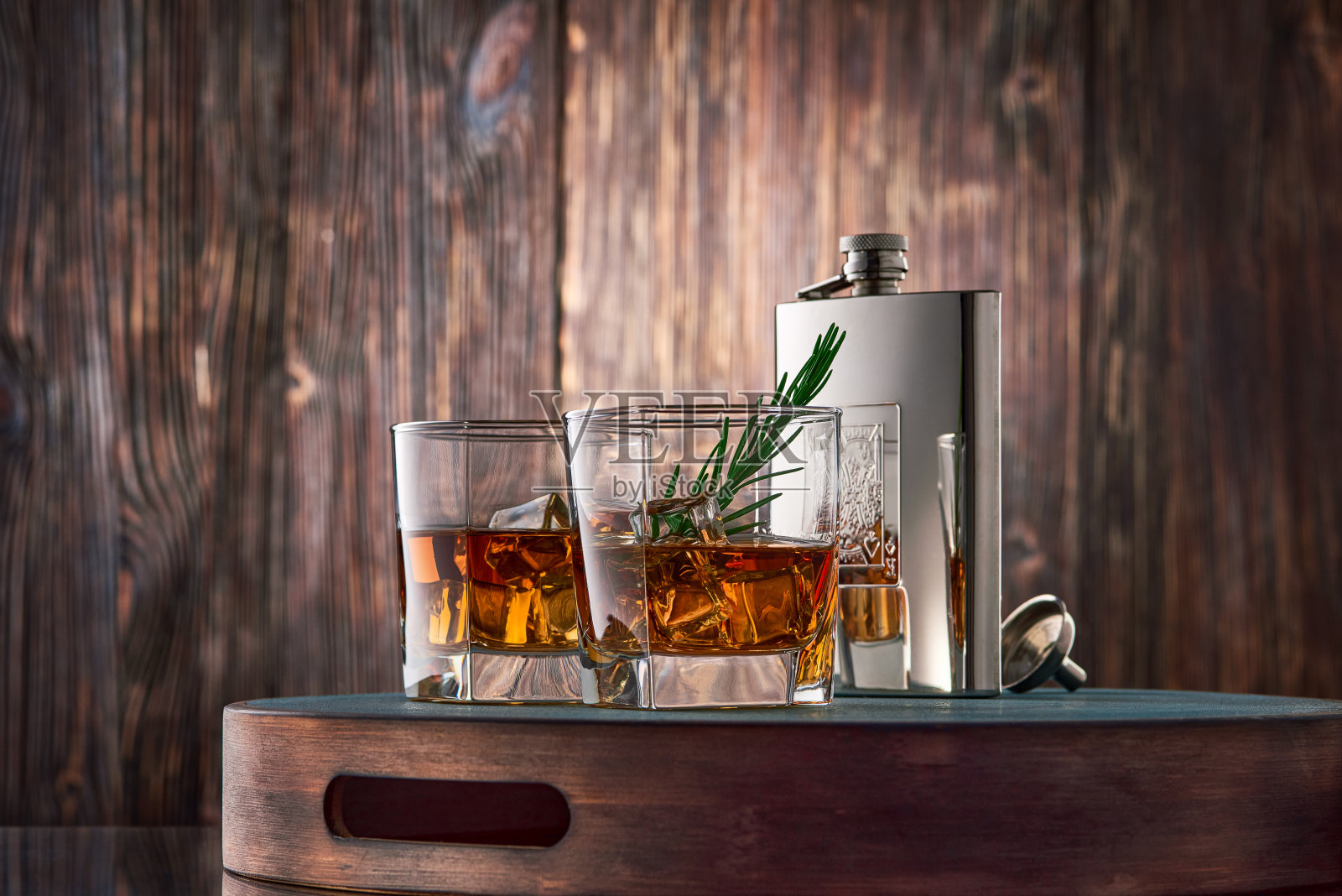 靠着一堵旧木墙的托盘上放着两杯加冰、一小枝迷迭香的方形威士忌和一只金属烧瓶。照片摄影图片
