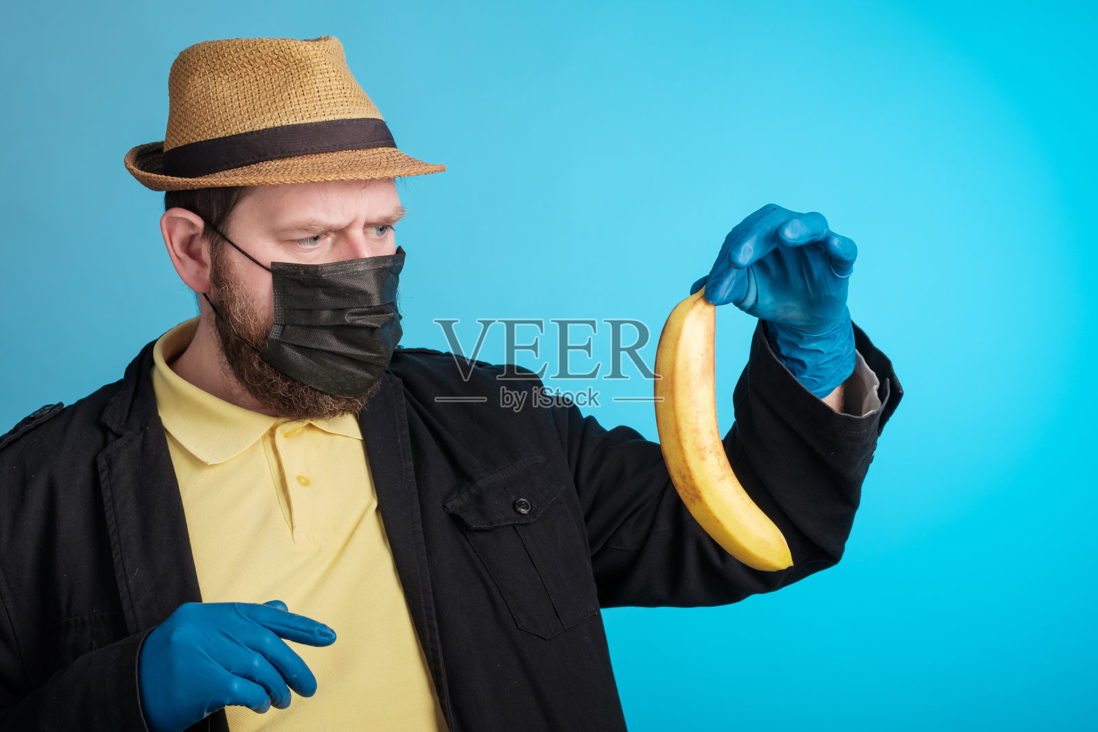 一个戴着面具和手套的男人怀疑地拿着一根香蕉看着它照片摄影图片