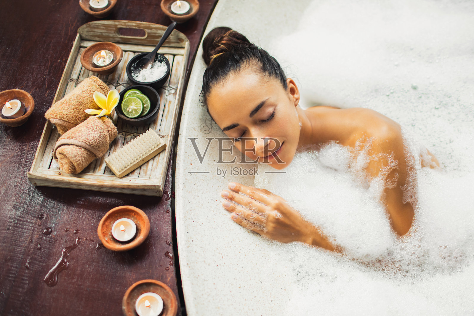 在浴缸中放松的女人。充满泡沫的浴缸。浪漫水疗，身体护理。天然化妆品套装用盐磨砂，毛巾，刷子放在木制托盘上。照片摄影图片