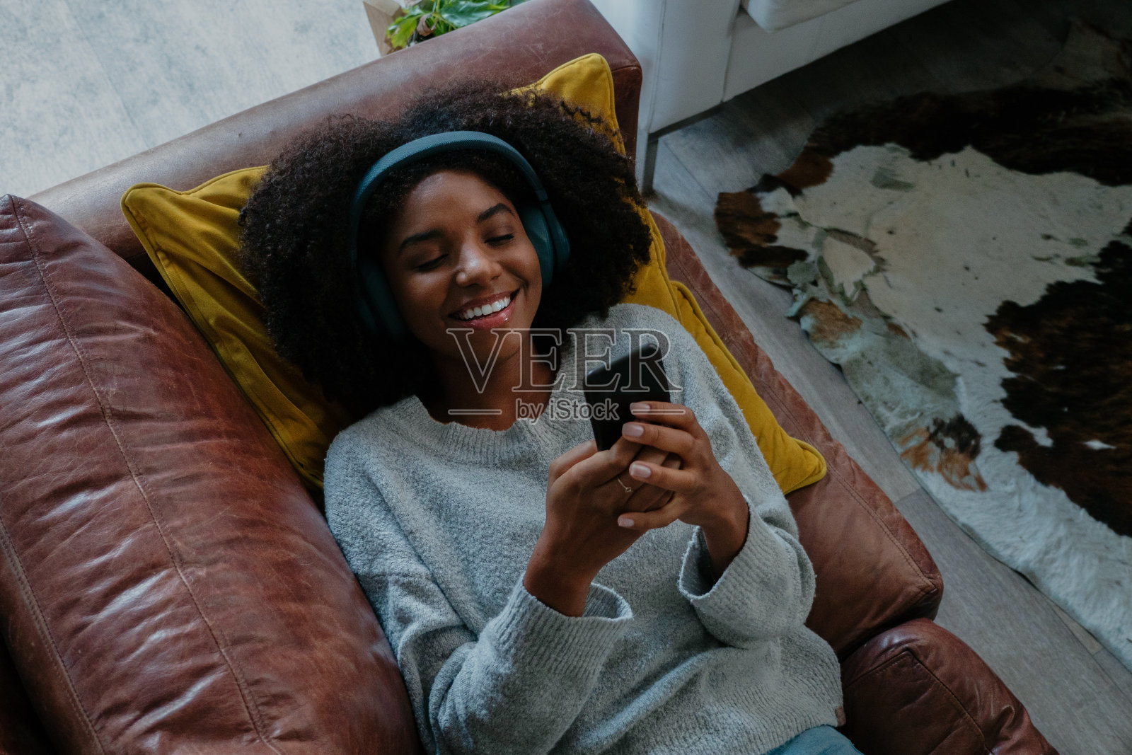 混血女性躺在沙发上微笑，而收到令人兴奋的短信在智能手机放松在现代休息室照片摄影图片