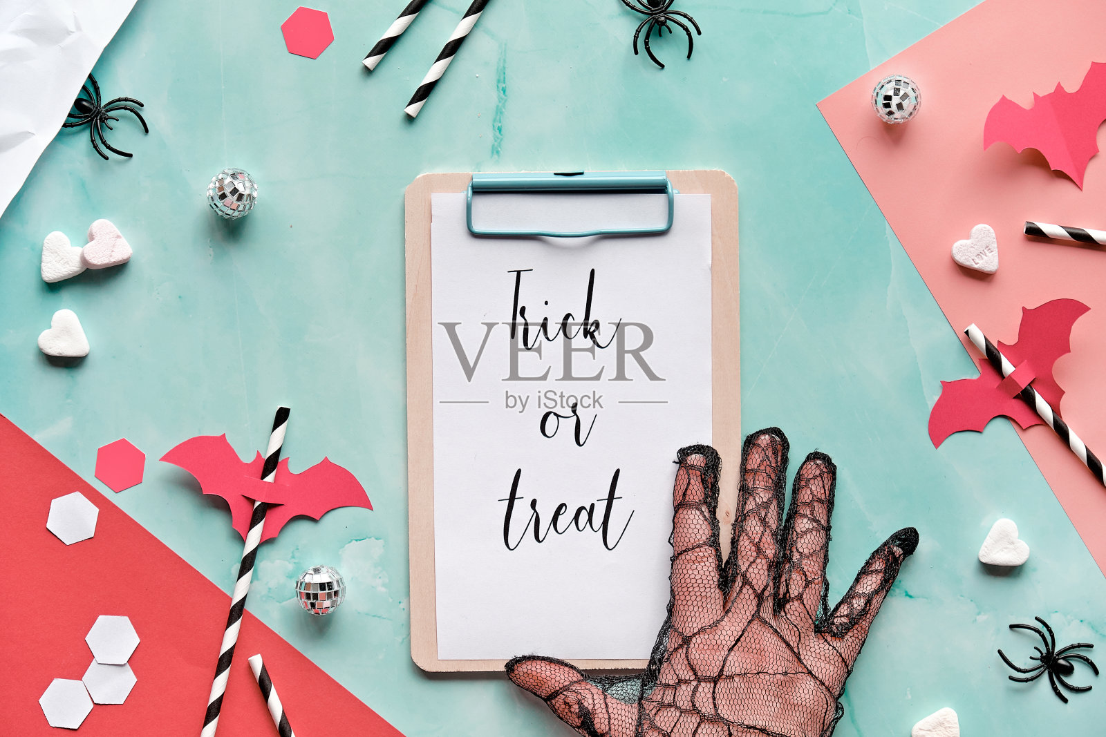 在剪贴板垫在薄荷绿和红色的分层纸背景上的Trick or treat文本。平铺，万圣节的安排与六边形的五彩纸屑，饮料吸管，蝙蝠剪影，糖心和蜘蛛。照片摄影图片