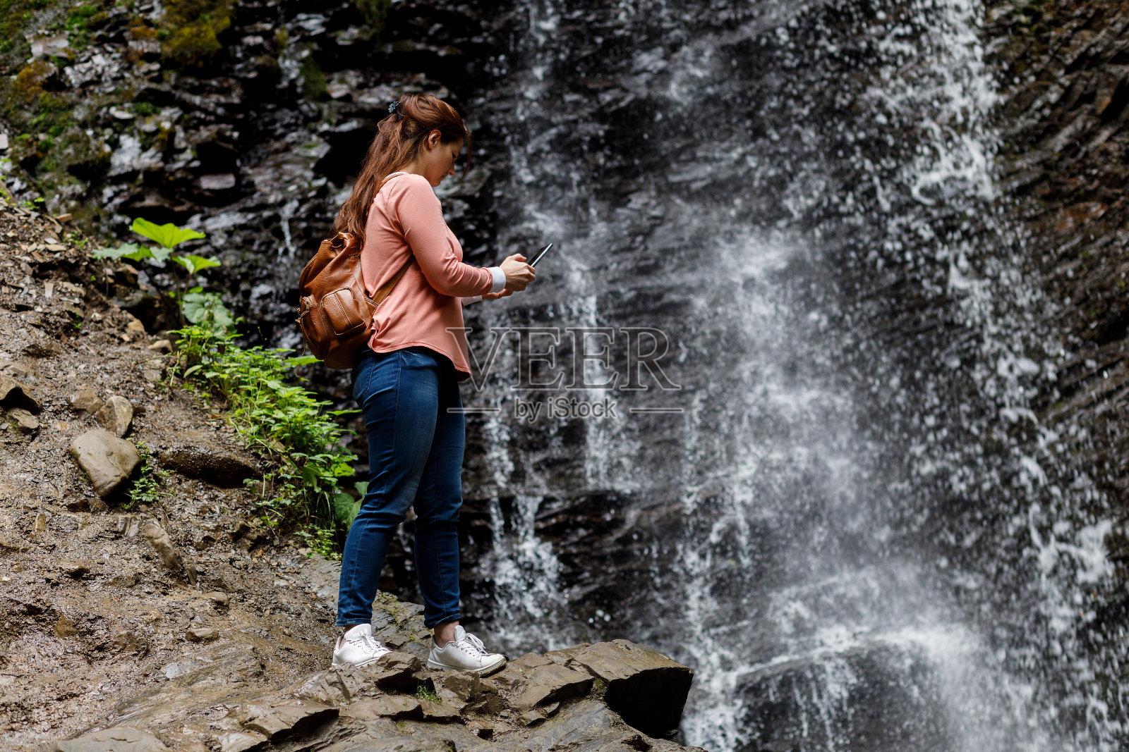 一个女人在一个瀑布的背景下用电话做布景。照片摄影图片