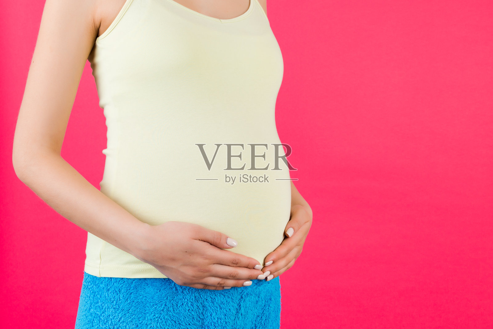 孕妇抱着她的腹部的裁剪图像。妈妈穿着色彩鲜艳的家装，背景是粉红色的。未来的父母期待着她的孩子。本空间照片摄影图片