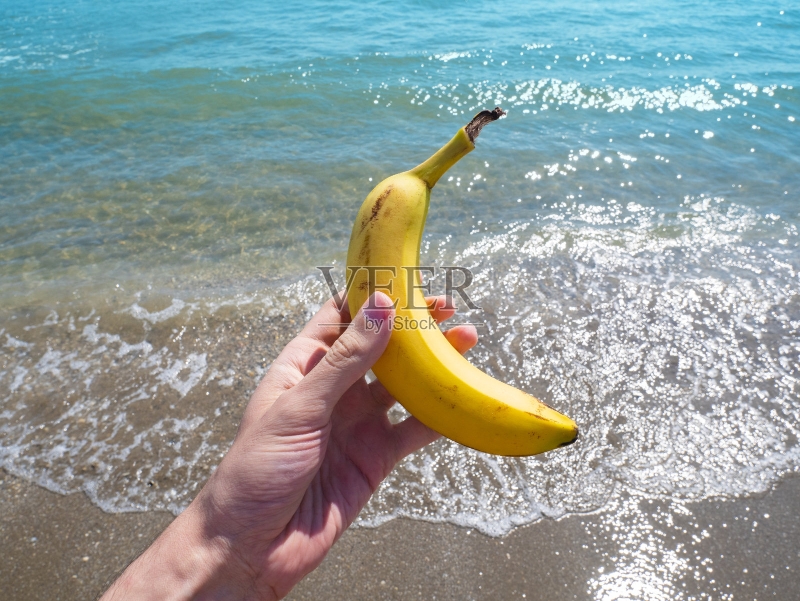 手里拿着香蕉。一只熟透了的黄香蕉，背景是沙滩上的海浪。健康的零食的概念。有趣的设计。照片摄影图片