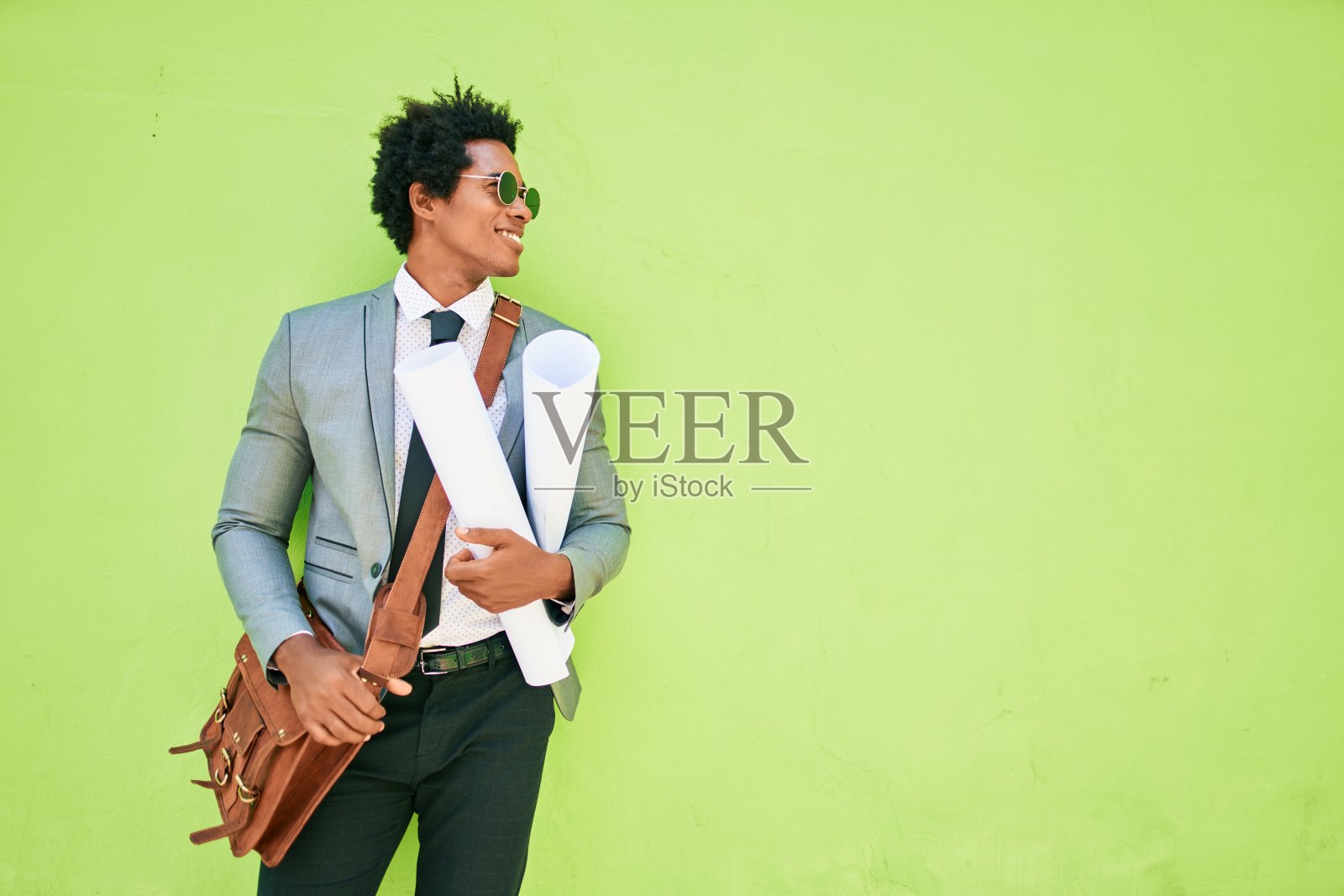 年轻英俊的非裔美国建筑师快乐的微笑。面带微笑靠在墙上，拿着蓝图走在镇上的大街上。照片摄影图片