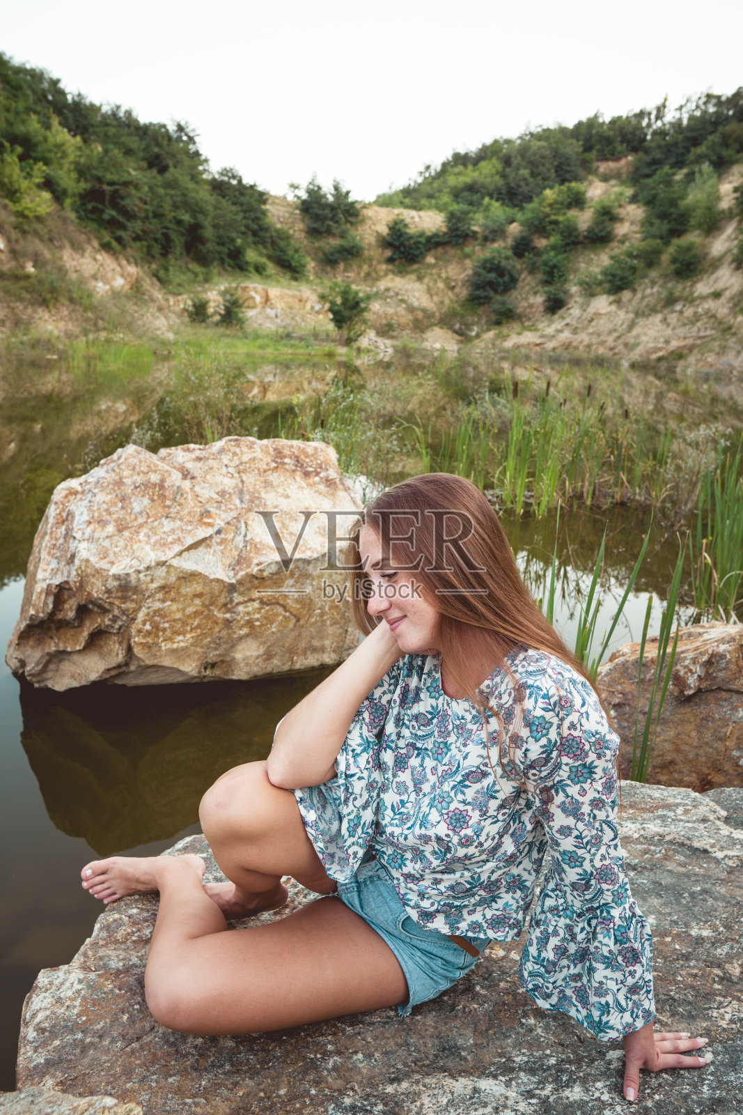 美丽年轻的姜黄色白人妇女坐在湖边。快乐的女人享受户外的自然。自由的概念。照片摄影图片