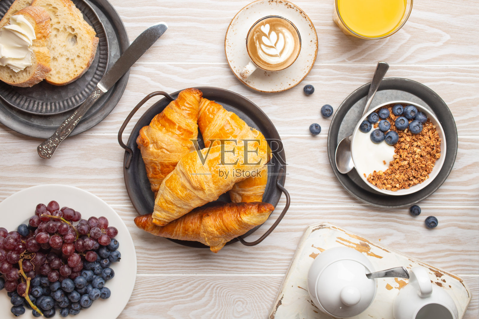 美丽的早餐，新鲜的羊角面包，格兰诺拉麦片，酸奶和浆果，面包，咖啡卡布奇诺和水果在盘子里。美味健康的早餐放在白色的木桌上，俯视图照片摄影图片