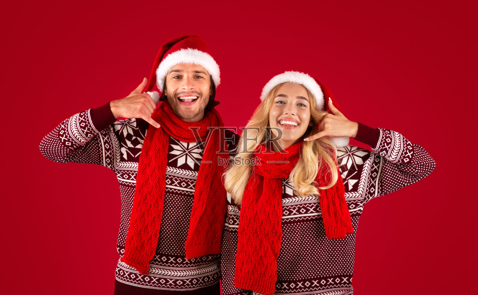 一对快乐的年轻夫妇在圣诞毛衣和圣诞老人帽显示叫我的手势在红色的背景照片摄影图片
