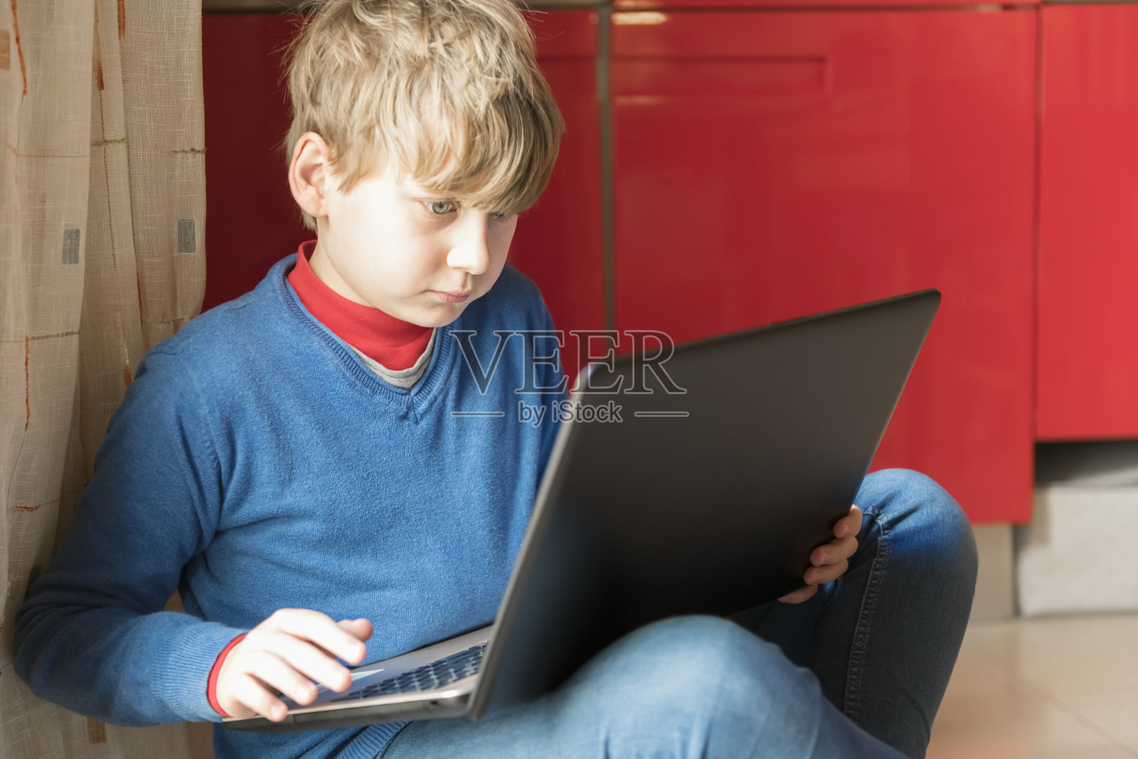 一个金发男孩正坐在房间的地板上，手里拿着笔记本电脑。在家教育和在家在线教育照片摄影图片