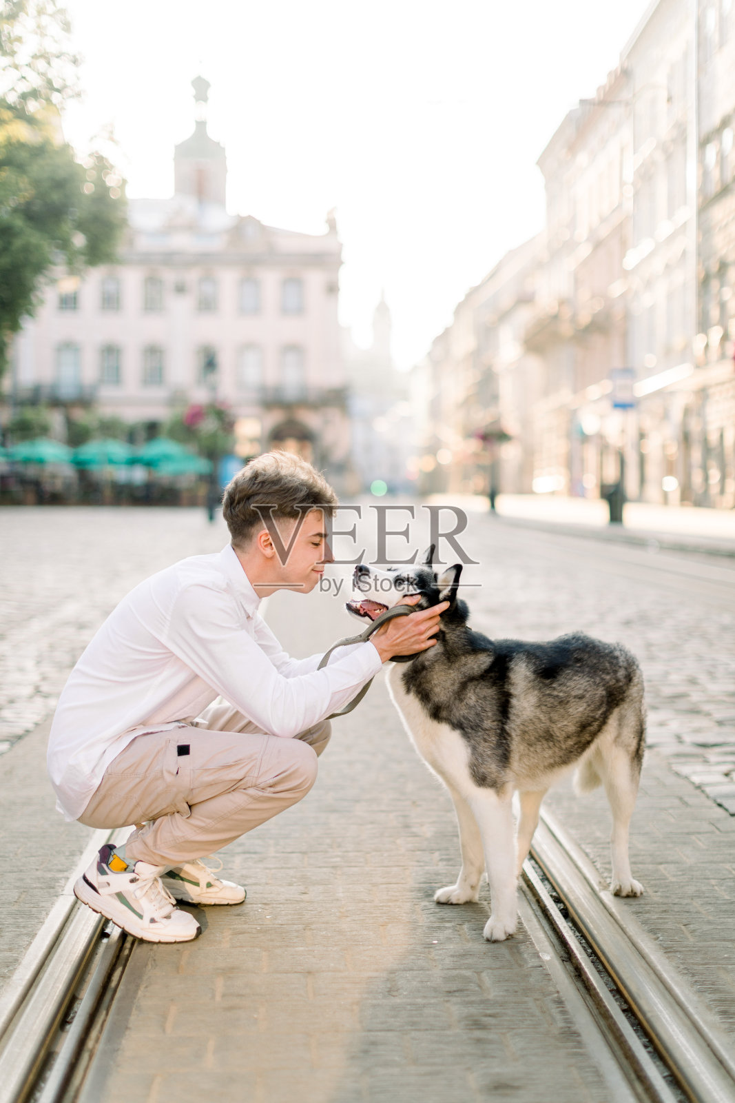 城市遛狗。年轻英俊的男孩穿着休闲装，在日出时分，带着他的哈士奇在古老的欧洲城市街道的人行道和电车轨道上散步照片摄影图片