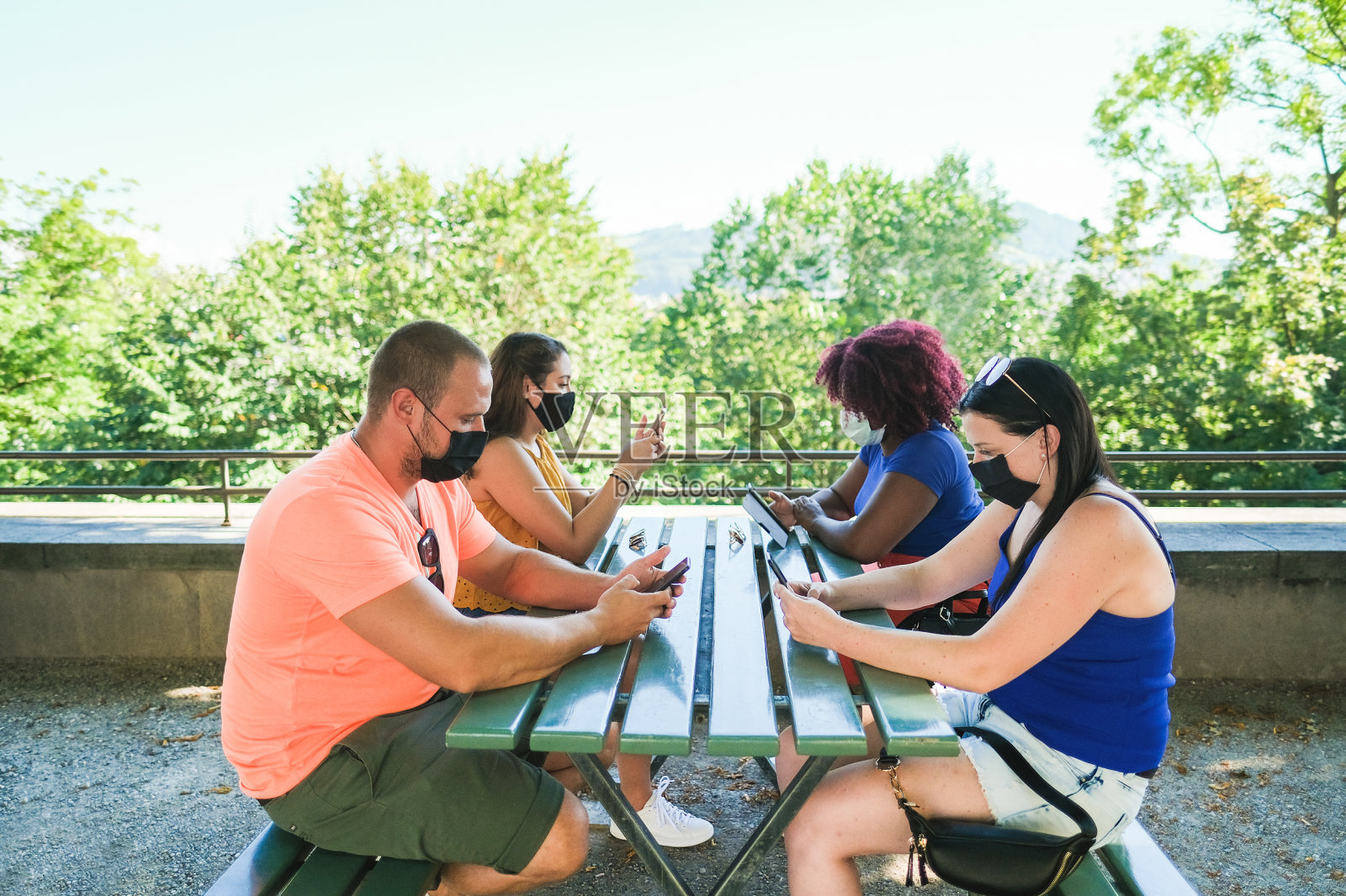 多种族朋友戴着防护口罩坐在长椅上看手机——冠状病毒生活方式照片摄影图片