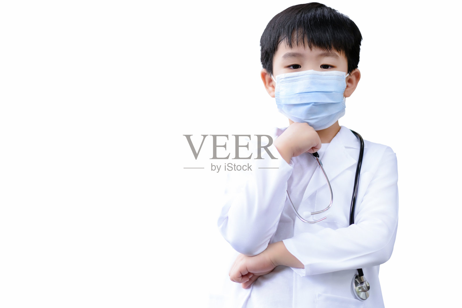 亚洲男孩穿着医疗服和防护卫生口罩，并将医生听诊器放在他的颈部隔离在白色背景。一个男孩抱着胳膊，摸着下巴，觉得很好玩。教育的概念。照片摄影图片