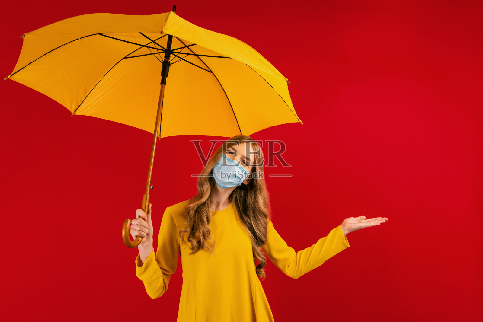一个开朗迷人的女孩穿着黄色的裙子，脸上戴着医用防护口罩，手里拿着一把黄色的雨伞，在一个孤立的红色背景上，概念的秋天，秋天的心情照片摄影图片