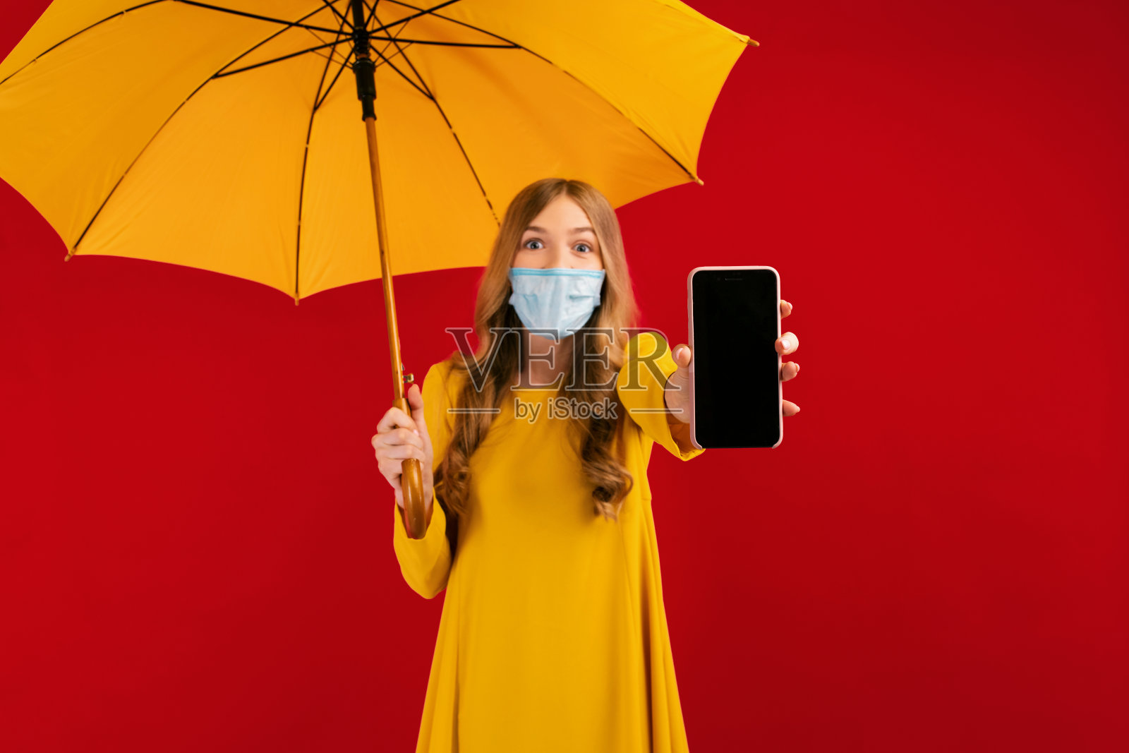 一个震惊的女孩，脸上戴着外科手术防护口罩，穿着黄色的裙子，手里拿着一把黄色的雨伞，展示着一个空的手机屏幕，在一个孤立的红色背景上复制空间照片摄影图片