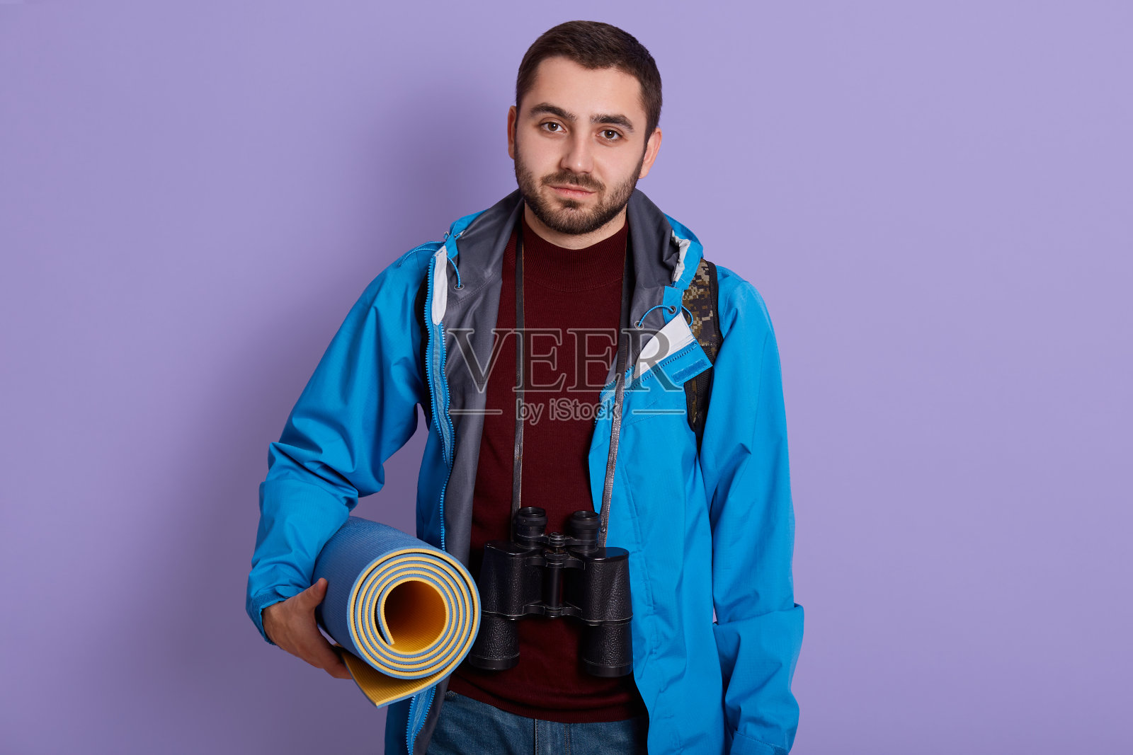 留着胡子的年轻人穿着蓝色夹克，拿着卷起的睡袋，看着摄像机，去露营，靠在淡紫色的墙上摆姿势，看起来很疲倦，脖子上挂着双筒望远镜。照片摄影图片
