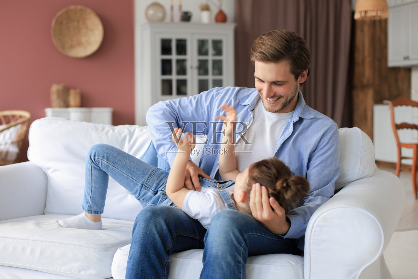 微笑的年轻父亲躺在客厅的沙发上和快乐的小女儿玩。照片摄影图片