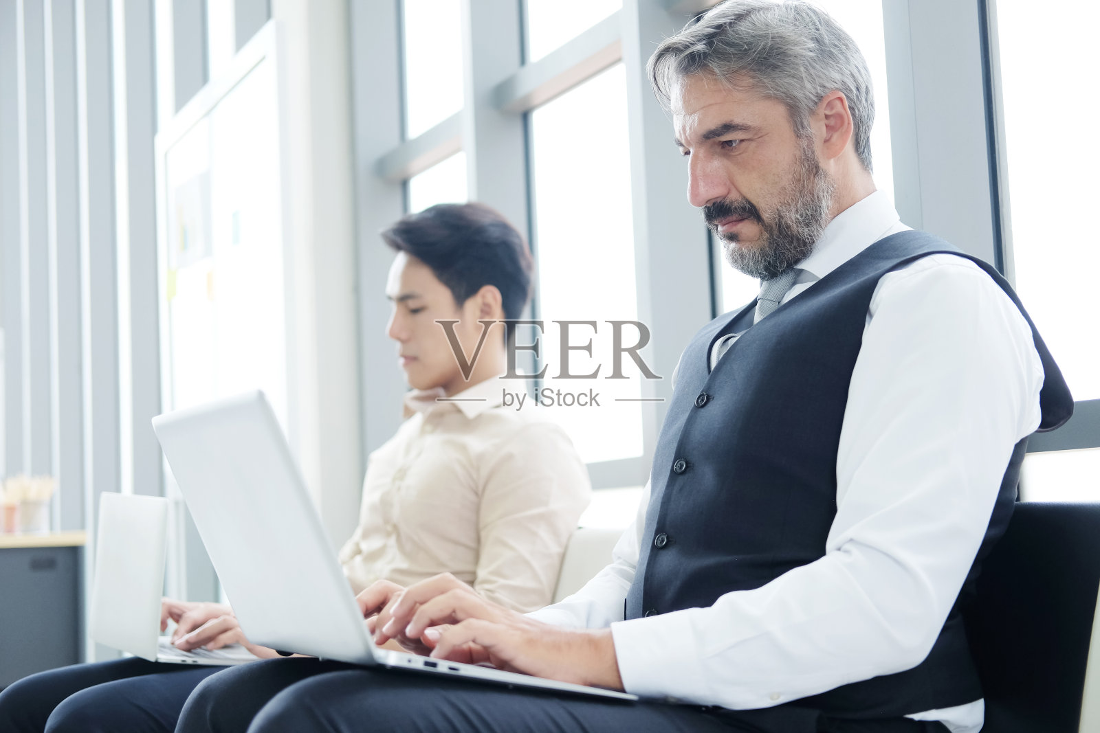 严肃的资深白种商人正在办公室工作与笔记本电脑。他与亚洲员工坐得距离远。技术为商业和新常态的生活理念。照片摄影图片