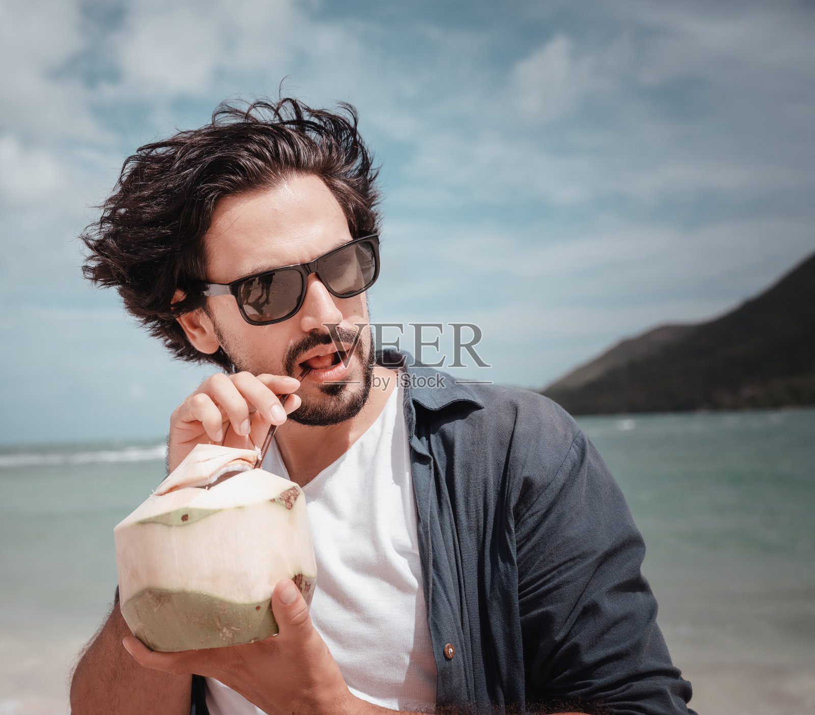 年轻英俊的拉丁美洲男人男人戴着太阳镜在热带海滩的热带海岸与新鲜的椰子在他的手照片摄影图片