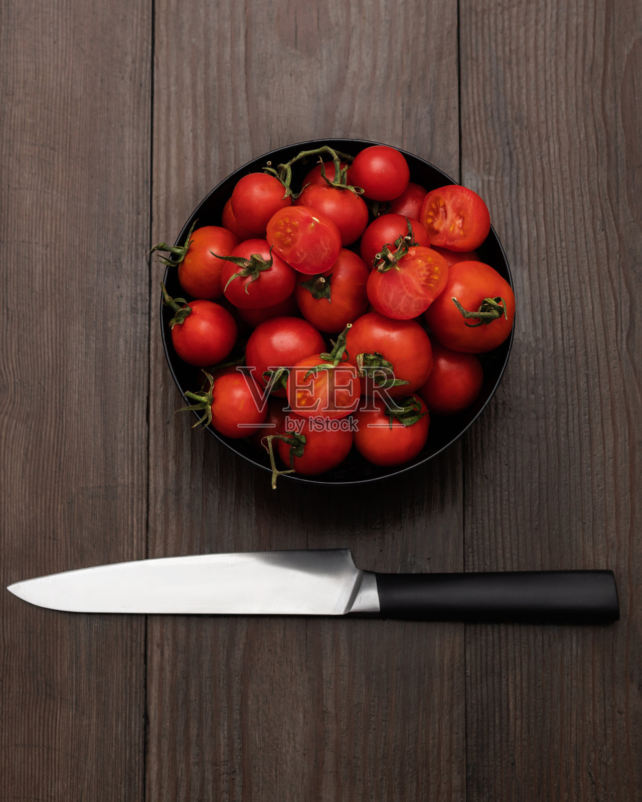 黑色木桌上放着黑色碗里的圣女果，旁边放着一把锋利的小刀。照片摄影图片