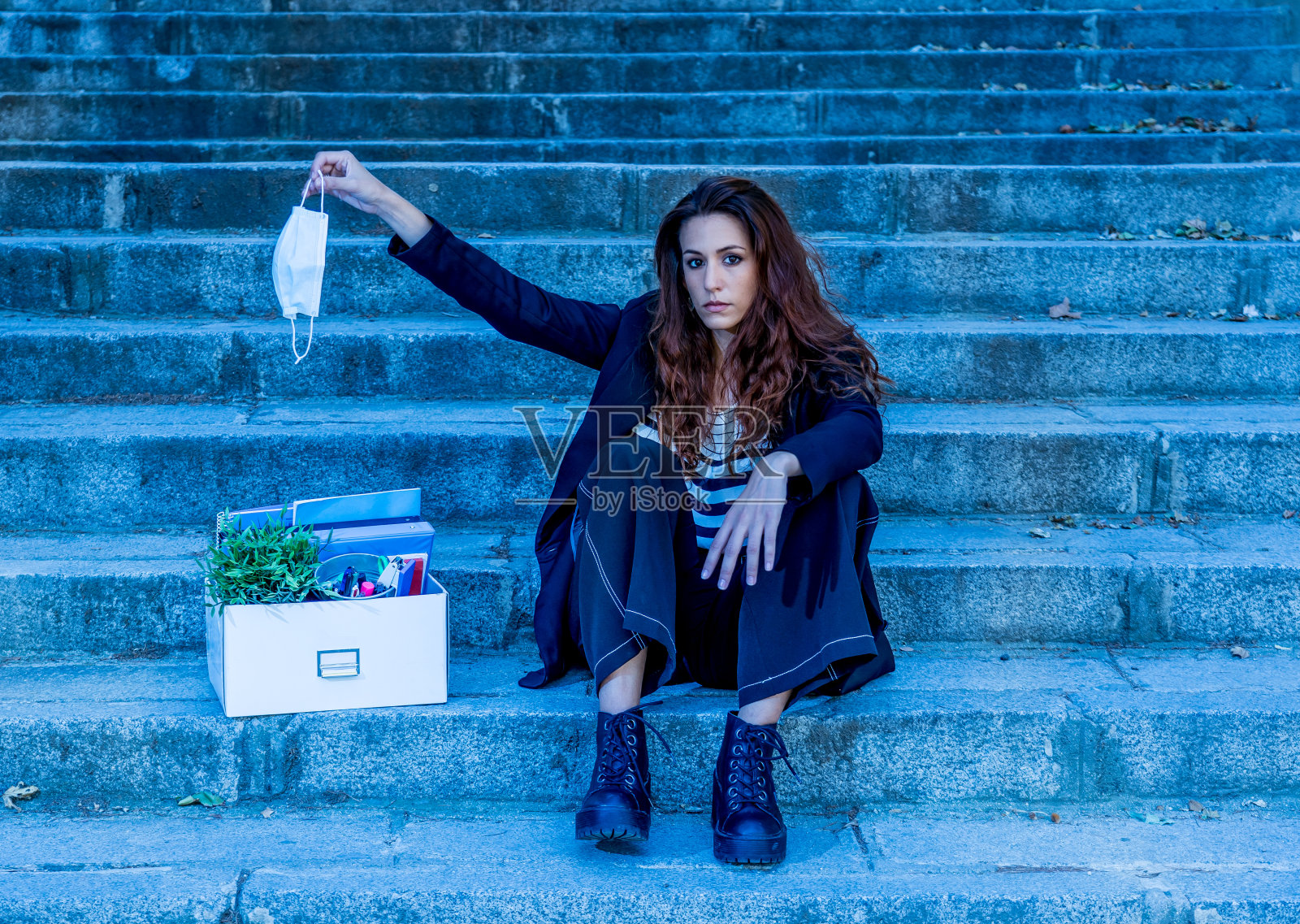 伤心被解雇的女人拿着口罩坐在楼梯上的商务区包厢里的办公人员。女性工人因冠状病毒裁员而失业。COVID-19经济影响的概念形象。照片摄影图片